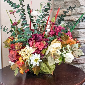 May include: A fall-themed floral arrangement in a square vase. The arrangement features red, white, and yellow flowers, along with green leaves and red berries.