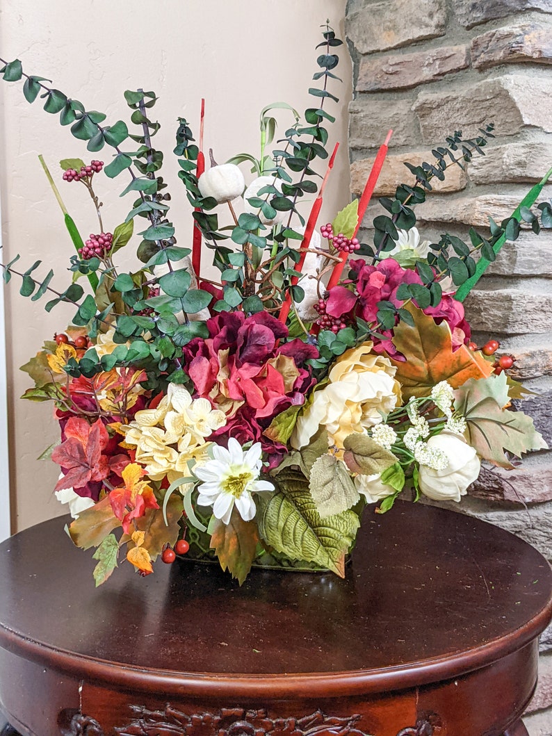 May include: A fall-themed floral arrangement in a square vase. The arrangement features red, white, and yellow flowers, along with green leaves and red berries.
