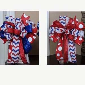 May include: Two red, white and blue ribbon bows with a chevron pattern, white polka dots and blue glitter. The bows are tied together and are on a white surface.