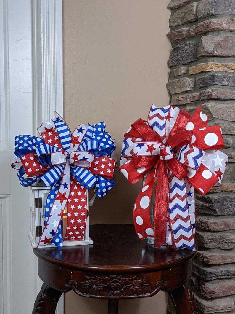 May include: Two red, white and blue ribbon bows with stars and polka dots. The bows are on a wooden table in front of a brick wall.