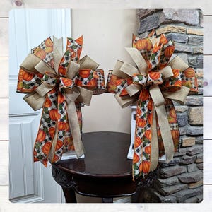 May include: Two decorative fall bows. Each bow features a combination of burlap, pumpkin-patterned, and black and white checkered ribbons. The bows are arranged on a dark brown table, with a white stand supporting each bow. The background includes a stone wall and a white door.