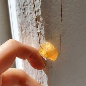 May include: A golden-yellow crystal pendant necklace. The raw, irregularly shaped crystal hangs from a delicate gold-colored chain. The necklace is displayed against a textured white wall, with sunlight casting shadows.