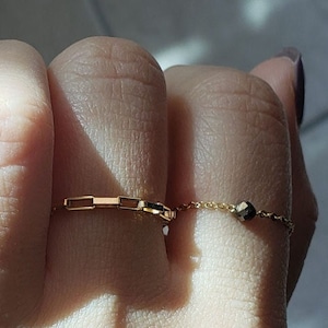 以下が含まれることがあります： 指に2つの金の指輪。上の指輪はチェーンリンクスタイルで、下の指輪は小さな丸いビーズが付いた繊細なチェーンです。