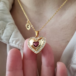 May include: Gold necklace with a heart pendant featuring a red rhinestone in the center. The pendant is surrounded by smaller rhinestones. The necklace also has a small charm with the letter "C" hanging from it.