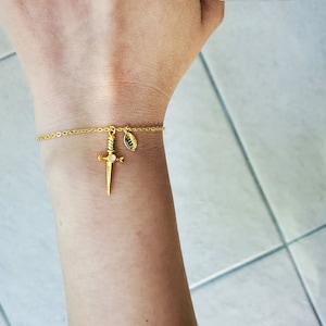 Puede incluir: Pulsera dorada con un dije de daga con una pequeña piedra blanca redonda. La pulsera también incluye un dije en forma de hoja con una inicial. Se lleva en la muñeca.