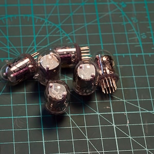 May include: A group of six silver vacuum tubes with glass casings and metal pins. The tubes are arranged in a cluster on a green cutting mat.