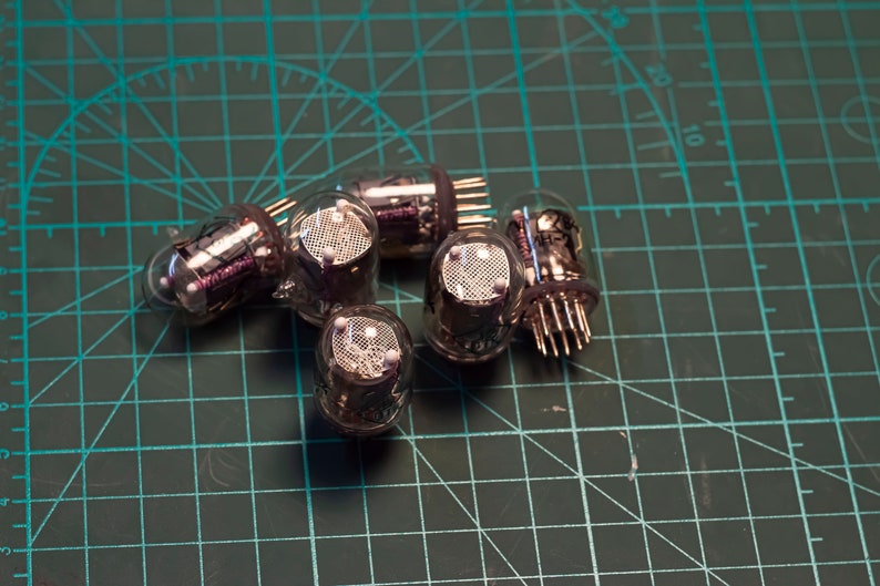 May include: A group of six silver vacuum tubes with glass casings and metal pins. The tubes are arranged in a cluster on a green cutting mat.