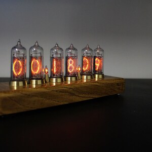May include: A wooden base with six clear glass Nixie tubes displaying the time as "00:18:09". The tubes are lit with an orange glow.
