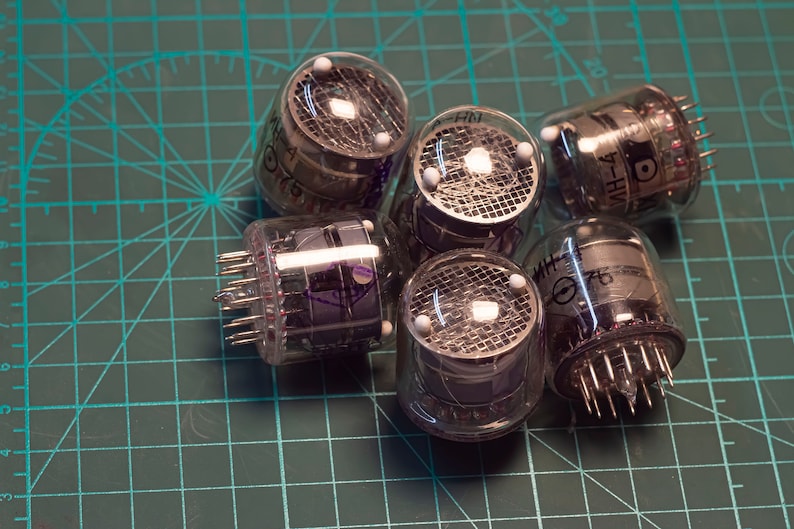 May include: A group of six clear glass vacuum tubes with metal grids and pins. The tubes have a purple tint and are labeled with the text "7-HVH" and "A-HM".