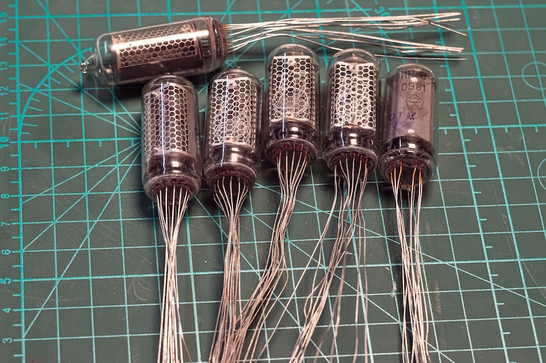 May include: Six vintage glass vacuum tubes with multiple wire leads extending from the bottom of each tube. The tubes are arranged in a row on a green grid surface.