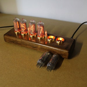 Puede incluir: Un reloj de madera con seis tubos Nixie naranjas brillantes que muestran la hora 15:47. Dos tubos Nixie de repuesto son visibles debajo del reloj.