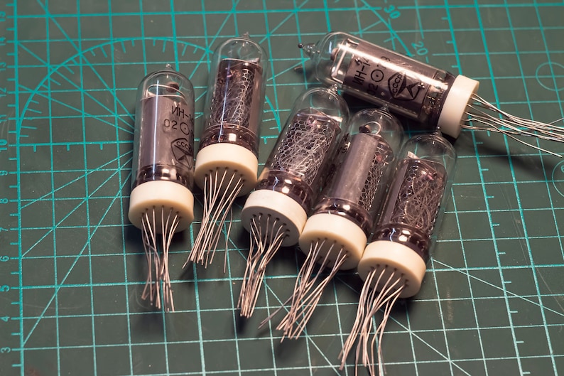 May include: Six vintage vacuum tubes with silver colored metal grids and clear glass bulbs. The tubes have white plastic bases with multiple pins extending from the bottom. The tubes are arranged on a green cutting mat.