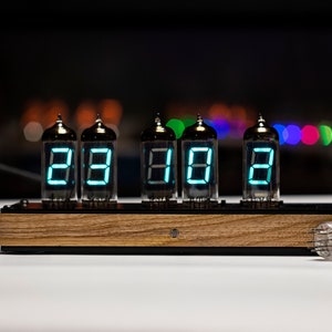 May include: A vintage-style clock with six Nixie tubes displaying the time as 23:18. The clock is made of wood and has a black metal frame. A spare Nixie tube is lying on the table next to the clock.