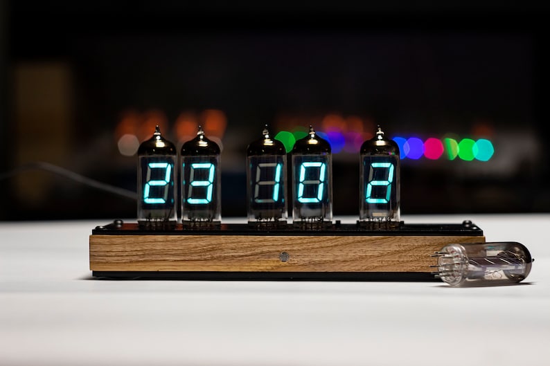 May include: A vintage-style clock with six Nixie tubes displaying the time as 23:18. The clock is made of wood and has a black metal frame. A spare Nixie tube is lying on the table next to the clock.