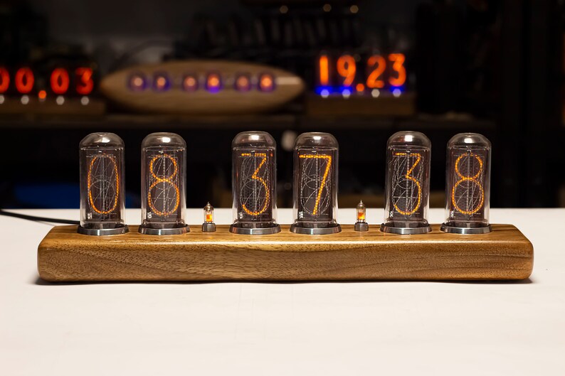 Puede incluir: Un reloj de madera con seis tubos Nixie naranjas que muestran la hora 08:37:38. El reloj est&aacute; sobre una superficie blanca.