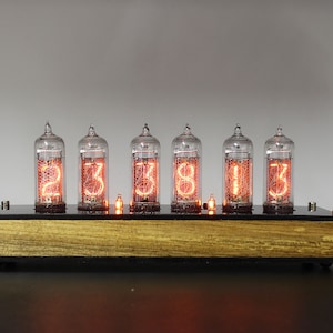 May include: A wooden base with six clear glass Nixie tubes displaying the time 23:38:13. The tubes are lit with a warm orange glow.