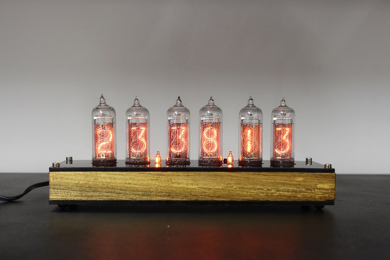May include: A wooden base with six clear glass Nixie tubes displaying the time 23:38:13. The tubes are lit with a warm orange glow.