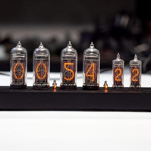 K&ouml;nnte beinhalten: Eine Digitaluhr mit sechs Glasr&ouml;hren, die die Zeit "00:54:22" anzeigt. Die Ziffern leuchten orangefarben in den R&ouml;hren. Die Uhr steht auf einem schwarzen Holzsockel mit kleinen orangefarbenen Lichtern.