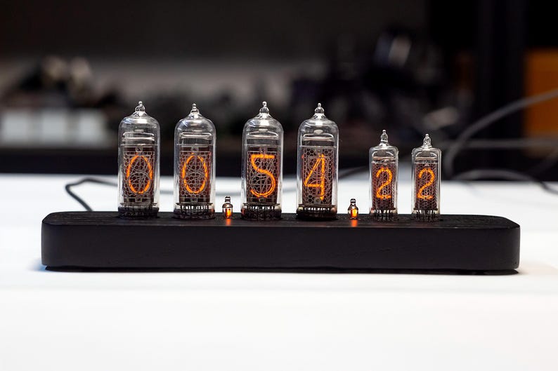 K&ouml;nnte beinhalten: Eine Digitaluhr mit sechs Glasr&ouml;hren, die die Zeit "00:54:22" anzeigt. Die Ziffern leuchten orangefarben in den R&ouml;hren. Die Uhr steht auf einem schwarzen Holzsockel mit kleinen orangefarbenen Lichtern.