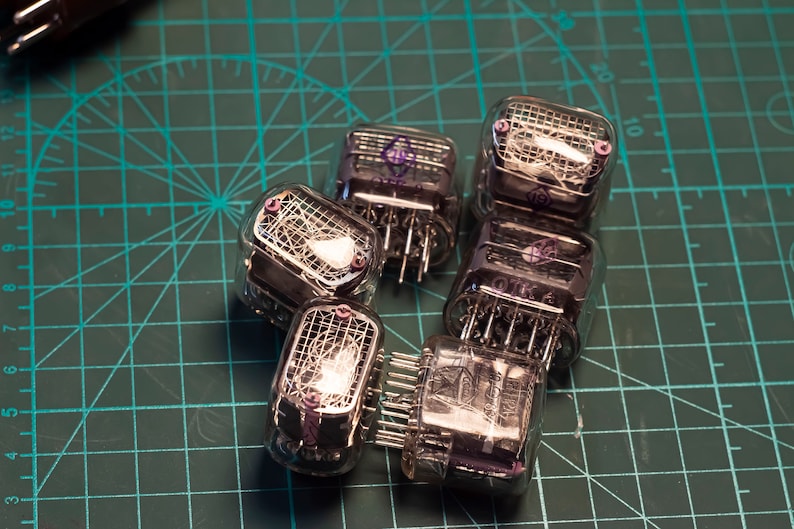 May include: A group of six clear glass Nixie tubes with glowing orange numbers inside. The tubes are arranged in a circle on a green cutting mat.