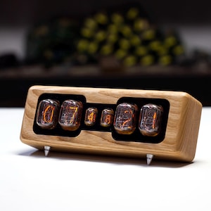 May include: A wooden digital clock with a light oak finish. The clock displays the time using glowing orange Nixie tubes. The clock has a rectangular shape and is supported by three silver metal feet.