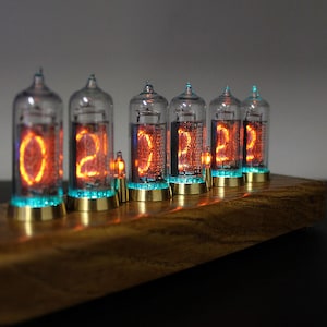 May include: A wooden clock with six glowing orange Nixie tubes displaying the time "02:22:22". The tubes are encased in glass and have a blue glow around the base.