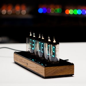 May include: A wooden clock with six Nixie tubes displaying the time in a bright blue-green colour. The clock is on a white surface.