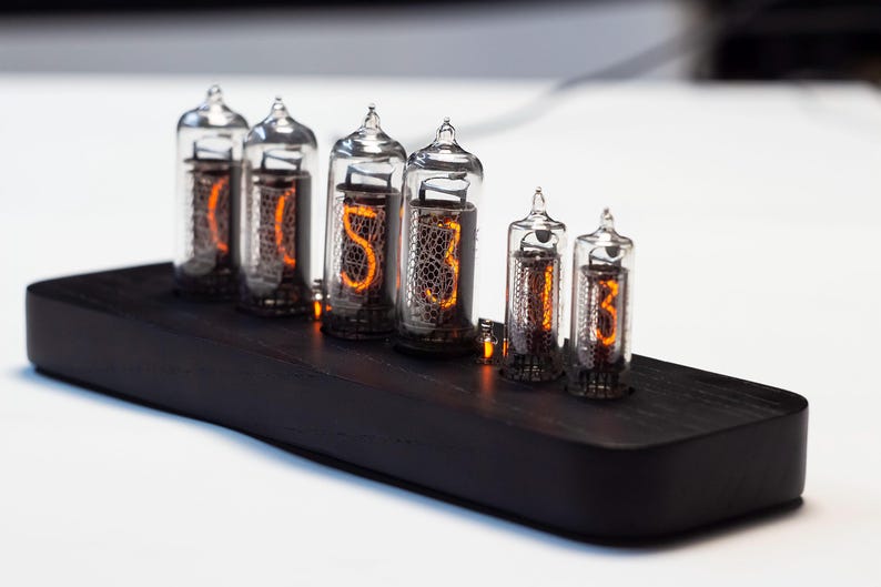K&ouml;nnte beinhalten: Eine Digitaluhr mit sechs Glasr&ouml;hren, die leuchtende orangefarbene Zahlen anzeigen. Die R&ouml;hren sind auf einem schwarzen Holzsockel montiert. Die Uhr hat ein Retro- und Industriedesign.