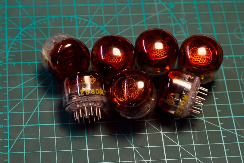 May include: A group of seven vintage vacuum tubes with orange glass bulbs and metal bases. The tubes are arranged in a semi-circle on a green cutting mat. The text "Z850M" is visible on the base of one of the tubes.