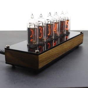 May include: A wooden and black clock with six clear glass Nixie tubes displaying the time as 12:34:56. The clock is plugged in with a black power cord.