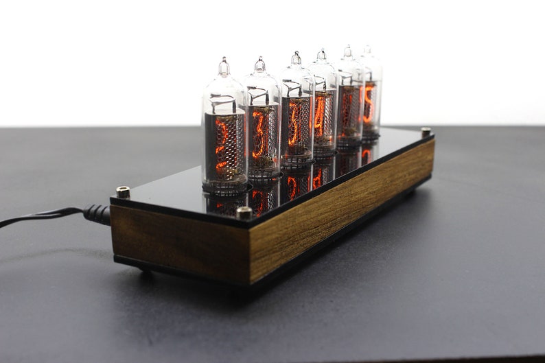 May include: A wooden and black clock with six clear glass Nixie tubes displaying the time as 12:34:56. The clock is plugged in with a black power cord.