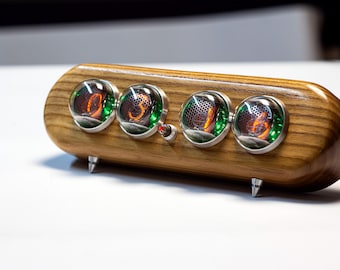 May include: A horizontal wooden clock featuring four circular nixie tubes with glowing orange digits and green internal lighting. The device is encased in a polished wood housing with silver metal accents and stands on two small silver metal feet.