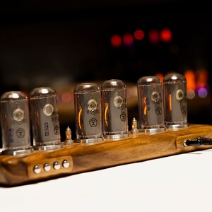 Puede incluir: Un reloj de madera con seis tubos Nixie brillantes que muestran la hora. El reloj est&aacute; alimentado por un cable negro.