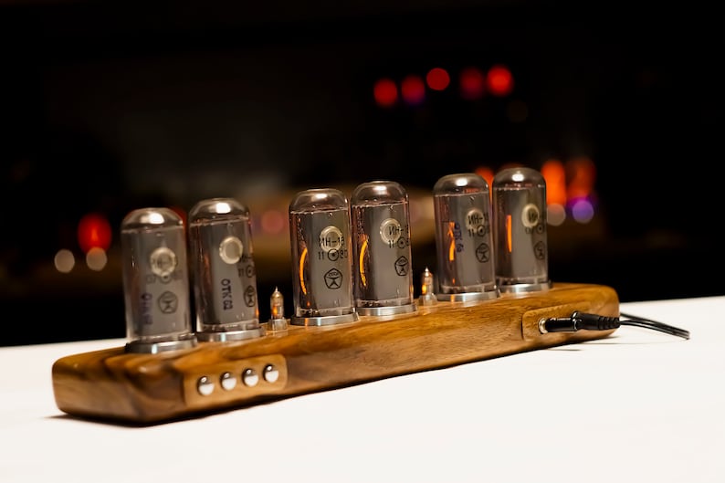 Puede incluir: Un reloj de madera con seis tubos Nixie brillantes que muestran la hora. El reloj est&aacute; alimentado por un cable negro.