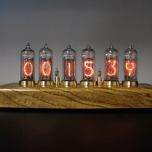 May include: A wooden clock with six glowing orange Nixie tubes displaying the time 001539. The clock has a brass base and is sitting on a black surface.
