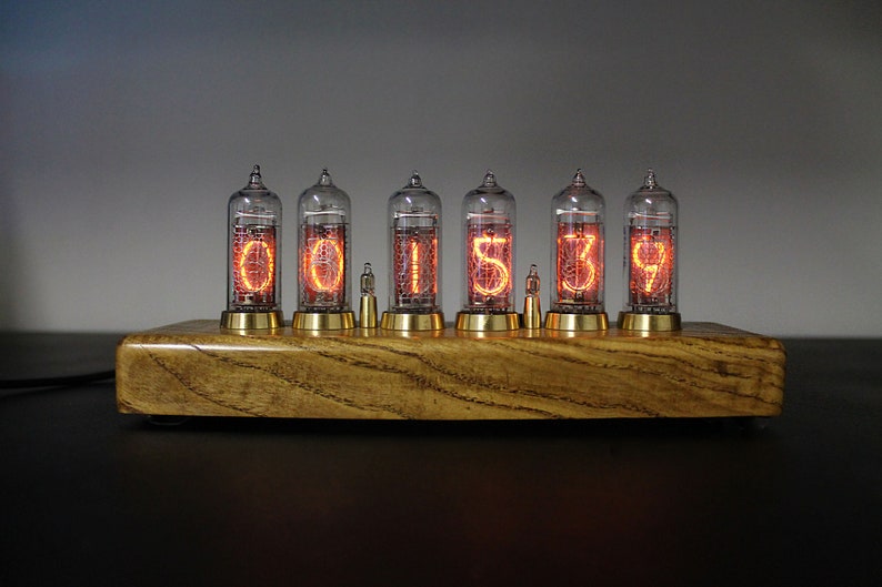May include: A wooden clock with six glowing orange Nixie tubes displaying the time 001539. The clock has a brass base and is sitting on a black surface.