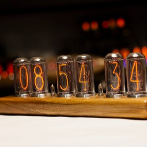 Puede incluir: Un reloj de madera con cuatro tubos Nixie que muestran la hora 08:54:34. Los tubos est&aacute;n iluminados con un brillo naranja.