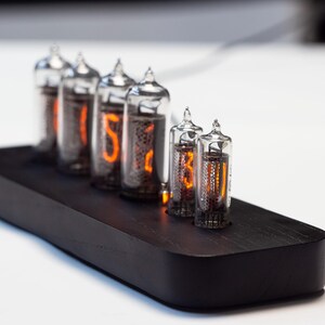 K&ouml;nnte beinhalten: Eine Digitaluhr mit sechs Glasr&ouml;hren, die orangefarbene Ziffern anzeigen. Die R&ouml;hren sind auf einem schwarzen Holzsockel montiert. Die Uhr ist ein elektronisches Ger&auml;t im Retro-Stil.
