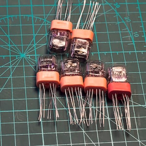 May include: Six vintage clear glass Nixie tubes with pink plastic bases and silver wires. The tubes are arranged in a triangular shape on a green grid surface.