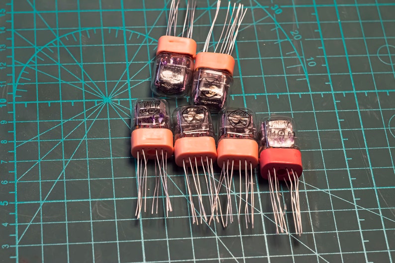 May include: Six vintage clear glass Nixie tubes with pink plastic bases and silver wires. The tubes are arranged in a triangular shape on a green grid surface.
