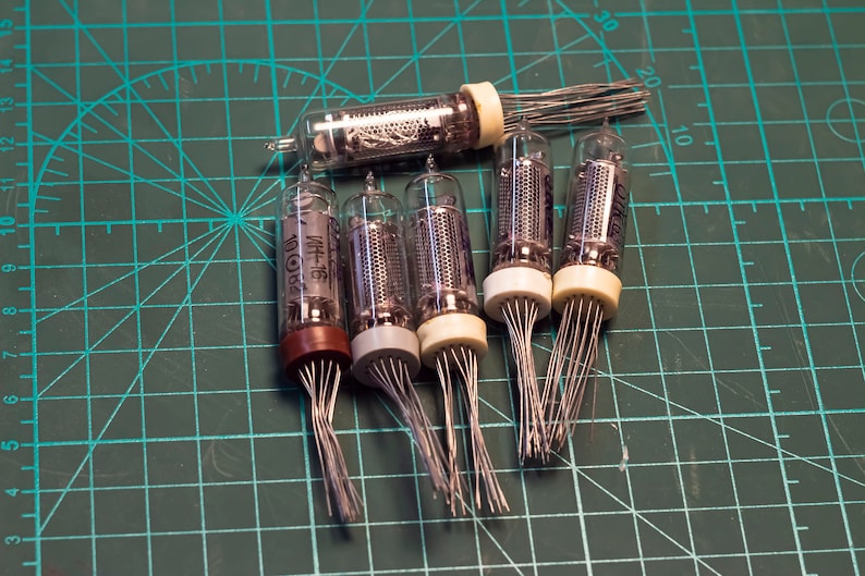 May include: Six vintage clear glass Nixie tubes with metal pins. The tubes have a grid pattern inside and are labeled with the text "ИН-16" in black. The tubes are arranged on a green cutting mat.