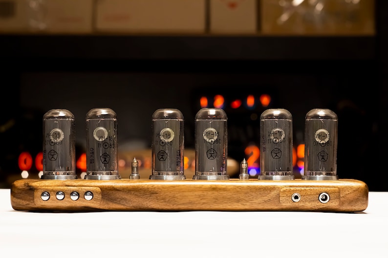 Puede incluir: Una base de madera con seis tubos de vac&iacute;o de vidrio transparente etiquetados "ИН-1Б 11 080". La base tiene dos conectores de audio en el lado derecho y cinco botones plateados en el lado izquierdo.