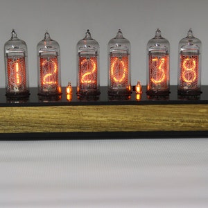 May include: A vintage-style clock with six clear glass tubes displaying the time as "12:03:38". The tubes are lit with a warm orange glow and are mounted on a black base with a wooden top.