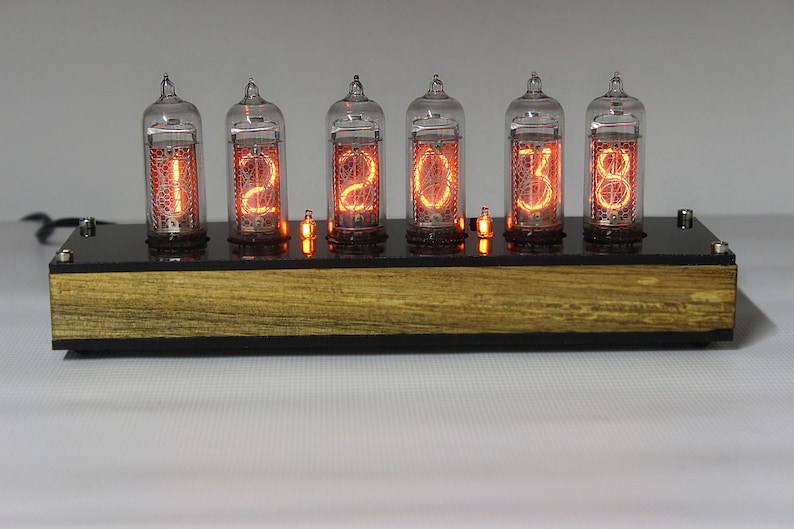 May include: A vintage-style clock with six clear glass tubes displaying the time as "12:03:38". The tubes are lit with a warm orange glow and are mounted on a black base with a wooden top.