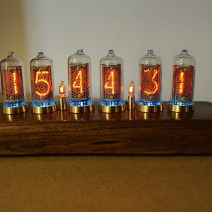 Puede incluir: Un reloj de madera con seis tubos Nixie que muestran la hora 15:44:31. Los tubos est&aacute;n iluminados con un brillo naranja y el reloj est&aacute; sentado sobre una base de madera.