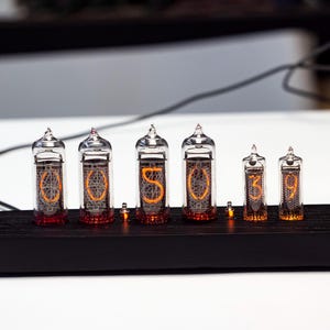 K&ouml;nnte beinhalten: Eine Digitaluhr mit sechs Glasr&ouml;hren, die die Uhrzeit "05:03:09" anzeigen. Die R&ouml;hren sind auf einem schwarzen Holzsockel montiert. Die Ziffern leuchten orange, und die Uhr hat eine Retro-Vintage-&Auml;sthetik.
