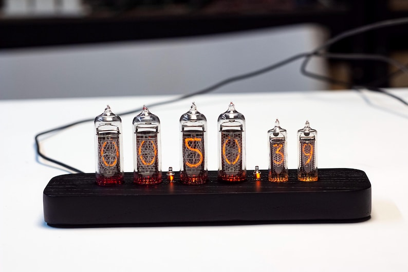 K&ouml;nnte beinhalten: Eine Digitaluhr mit sechs Glasr&ouml;hren, die die Uhrzeit "05:03:09" anzeigen. Die R&ouml;hren sind auf einem schwarzen Holzsockel montiert. Die Ziffern leuchten orange, und die Uhr hat eine Retro-Vintage-&Auml;sthetik.