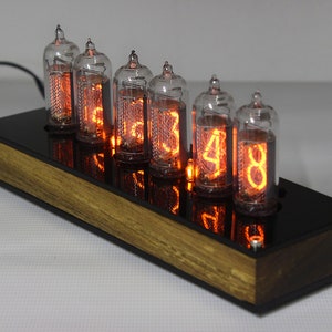 May include: A vintage-style clock with six orange glowing Nixie tubes displaying the time. The clock is mounted on a wooden base with a black acrylic face.