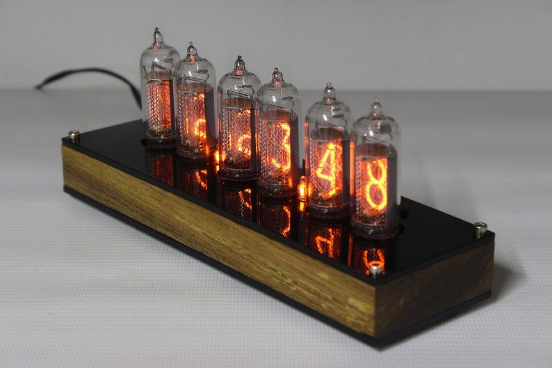 May include: A vintage-style clock with six orange glowing Nixie tubes displaying the time. The clock is mounted on a wooden base with a black acrylic face.