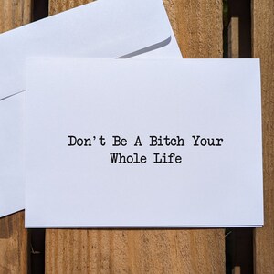 May include: White card with black text that reads "Don't Be A Bitch Your Whole Life" on a wooden background.